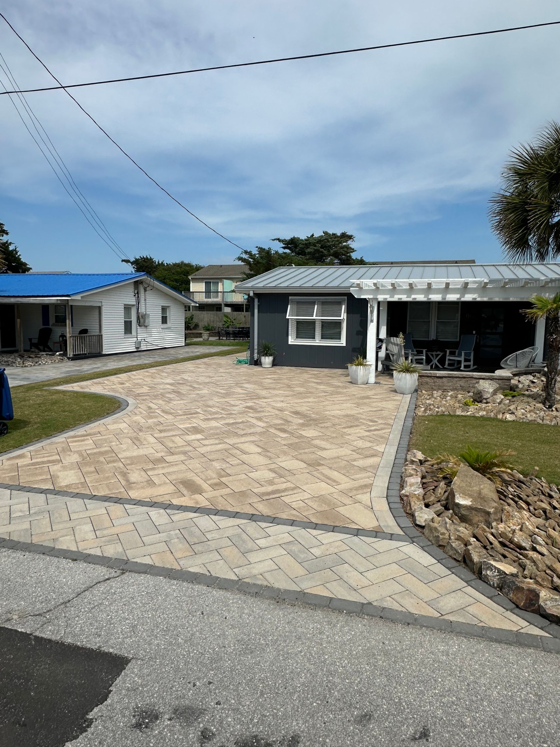 A beach house with a herringbone-patterned brick driveway. The house is painted dark gray with a white window frame.