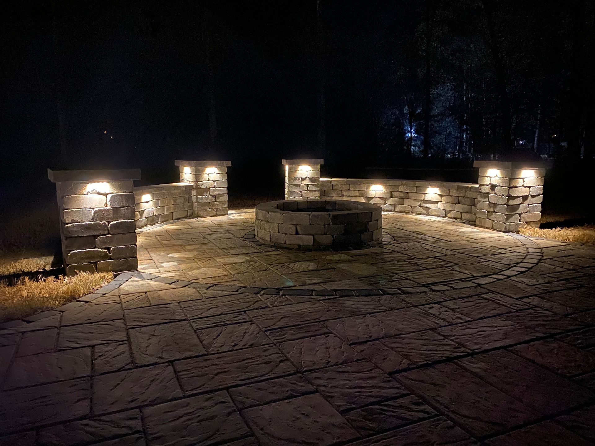 Night-lit stone fire pit area with built-in seating. Pillars and ground lit with warm lights, dark background.