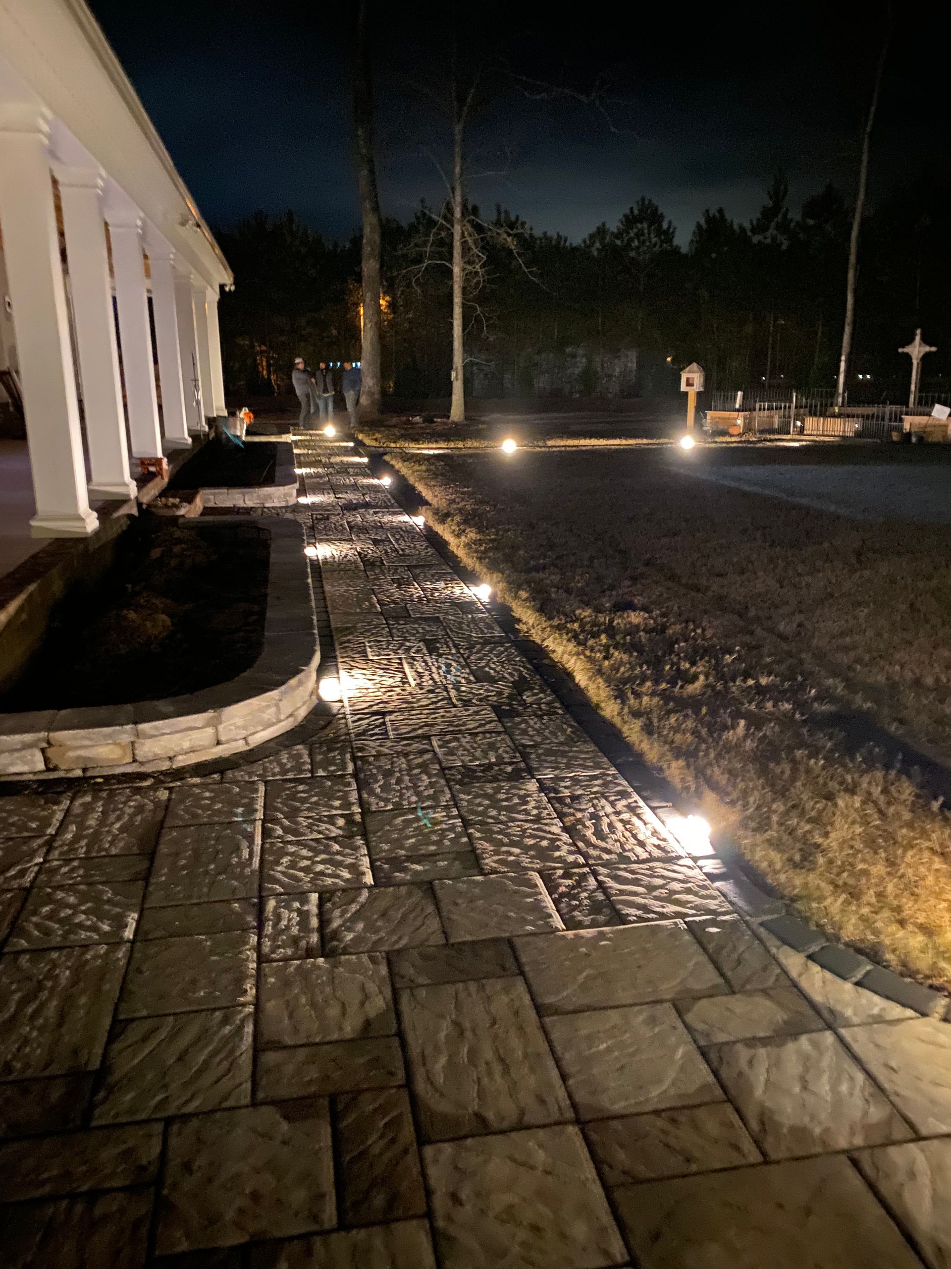 Brick walkway illuminated by ground lights at night, leading past a building with columns and a dark garden bed.
