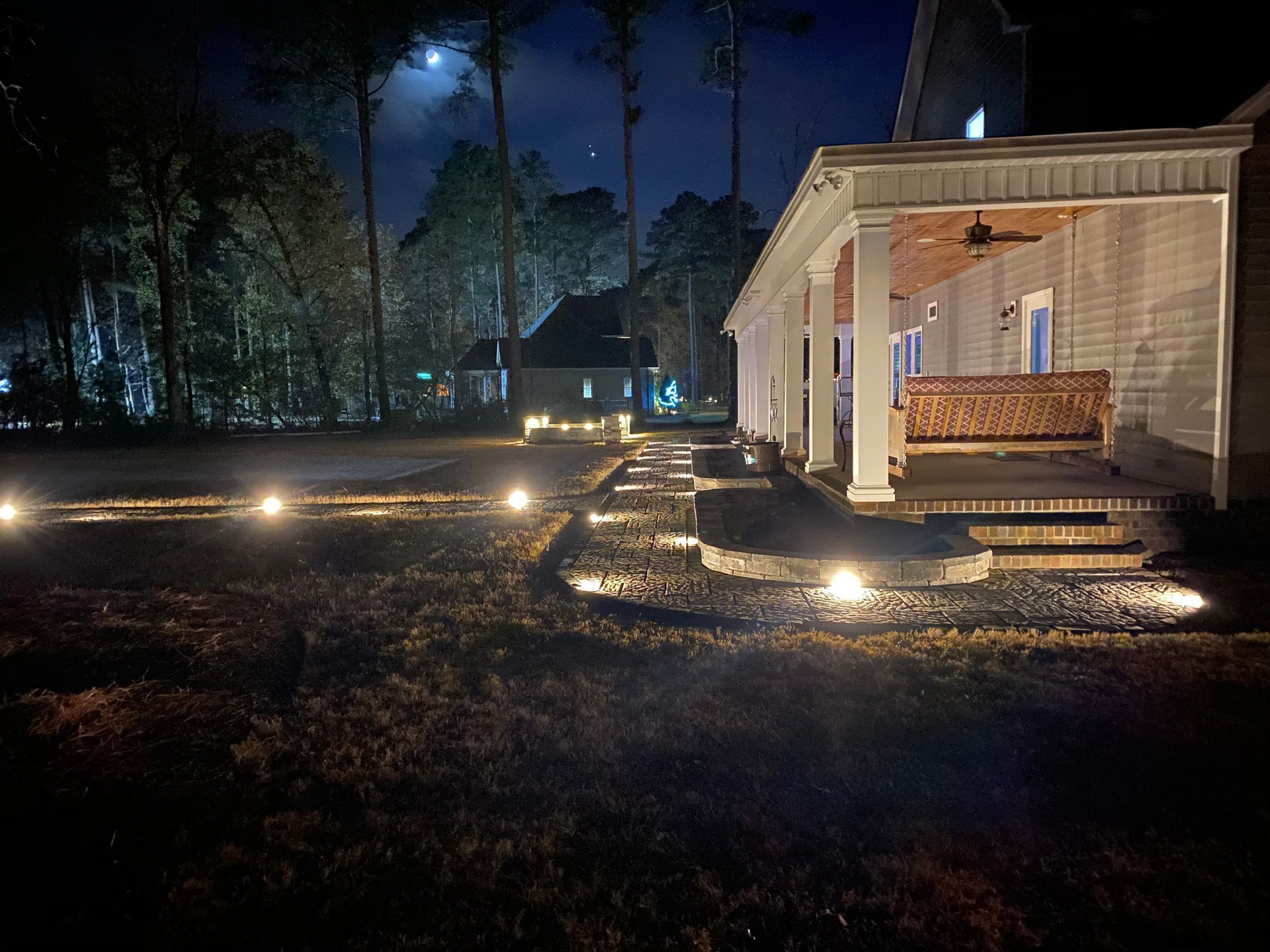 Nighttime view of a house with a lit pathway, porch, and a distant building; the moon is visible.