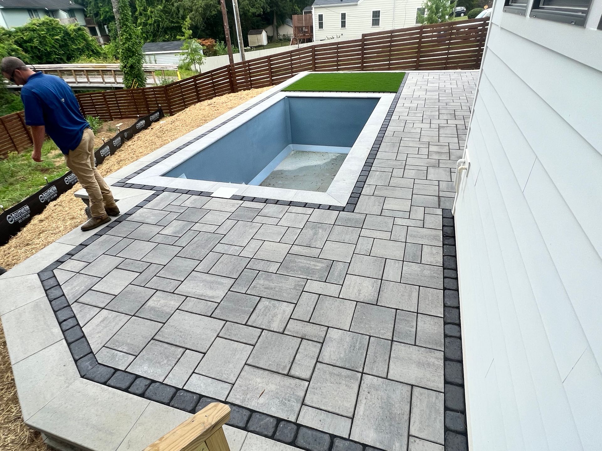 Backyard pool under construction, featuring gray pavers, retaining walls, and a person working.