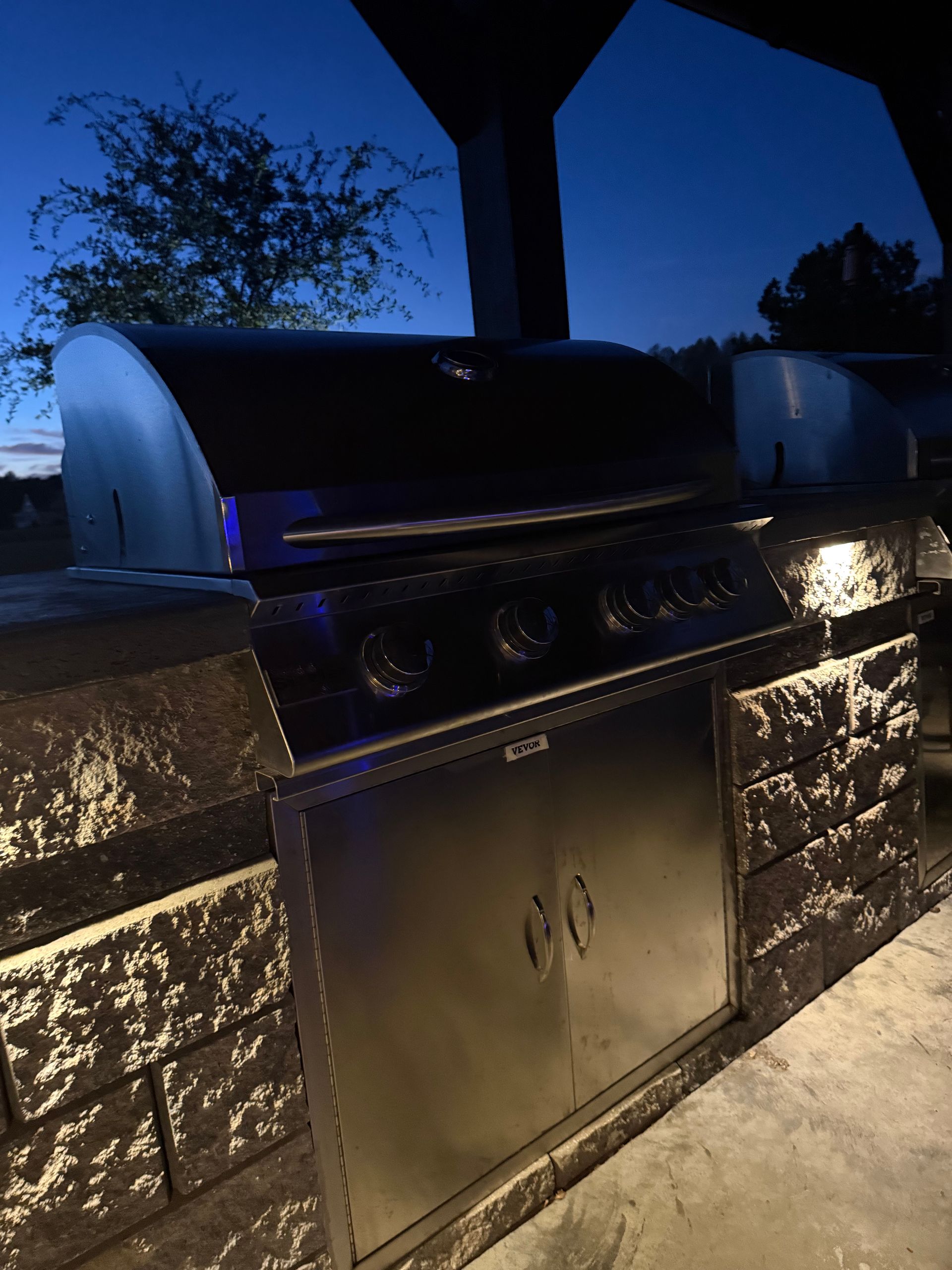 Outdoor grill built into a stone structure, lit at dusk. Stainless steel with four burners and storage.