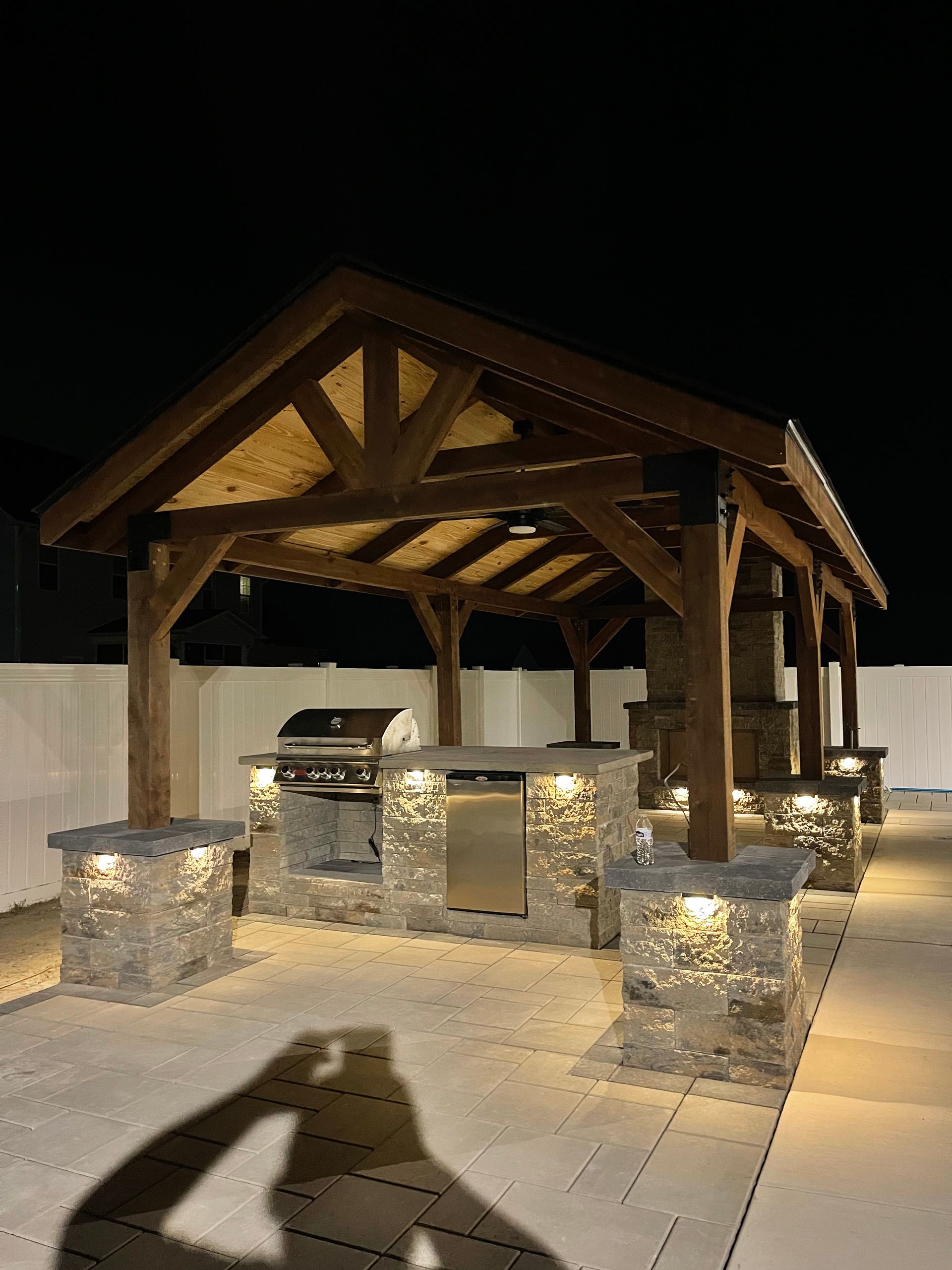 Outdoor kitchen under a wooden pergola at night, lit by spotlights.
