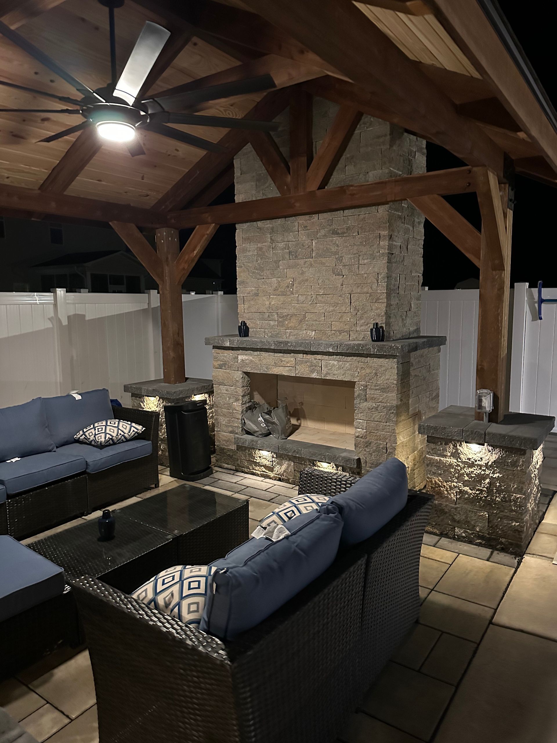 Outdoor patio with fireplace, seating, and wooden pergola. Blue couches, stone fireplace.
