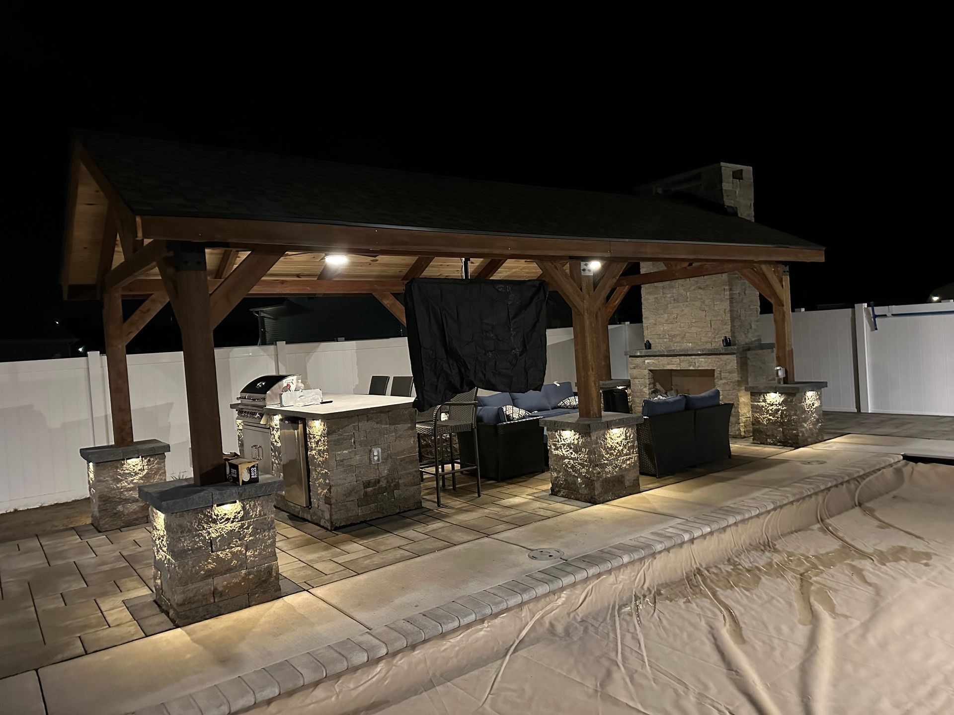 Outdoor patio with stone accents, a grill, and fireplace, under a wooden roof, at night.