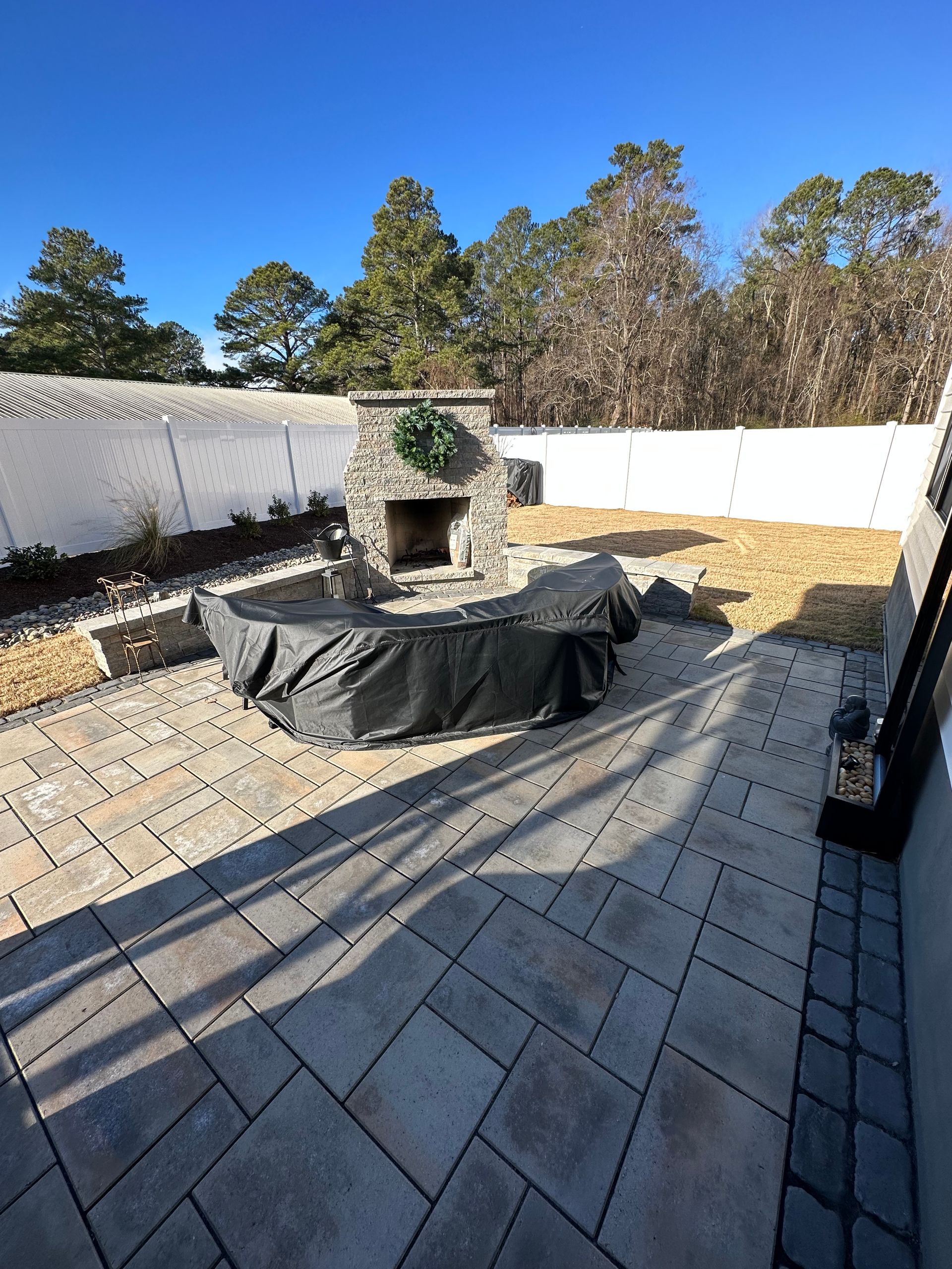 Outdoor patio with fireplace, paved area, covered seating, and a white fence under a blue sky.