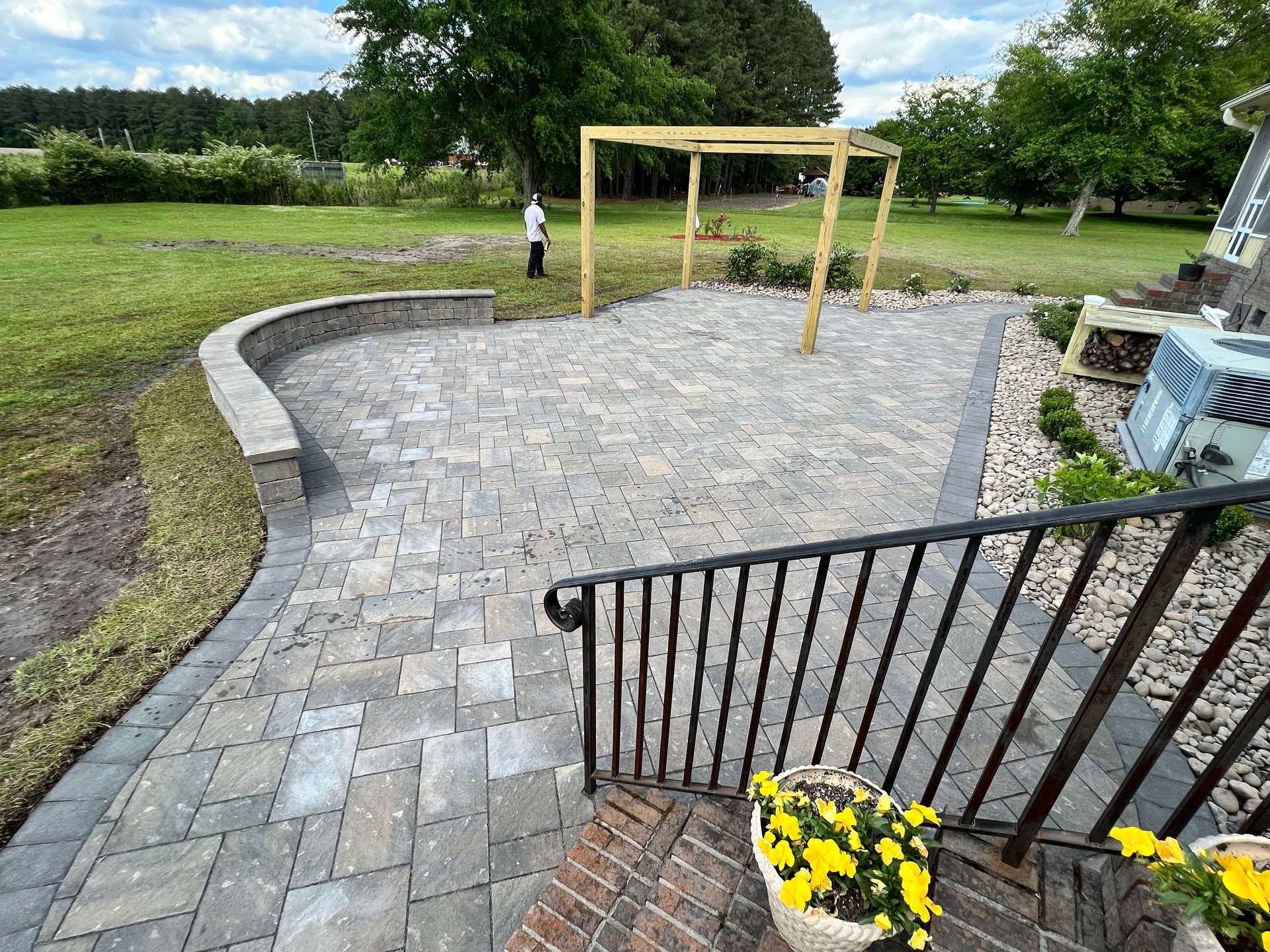 Brick patio with curved retaining wall and wooden pergola. Grass and trees in background.