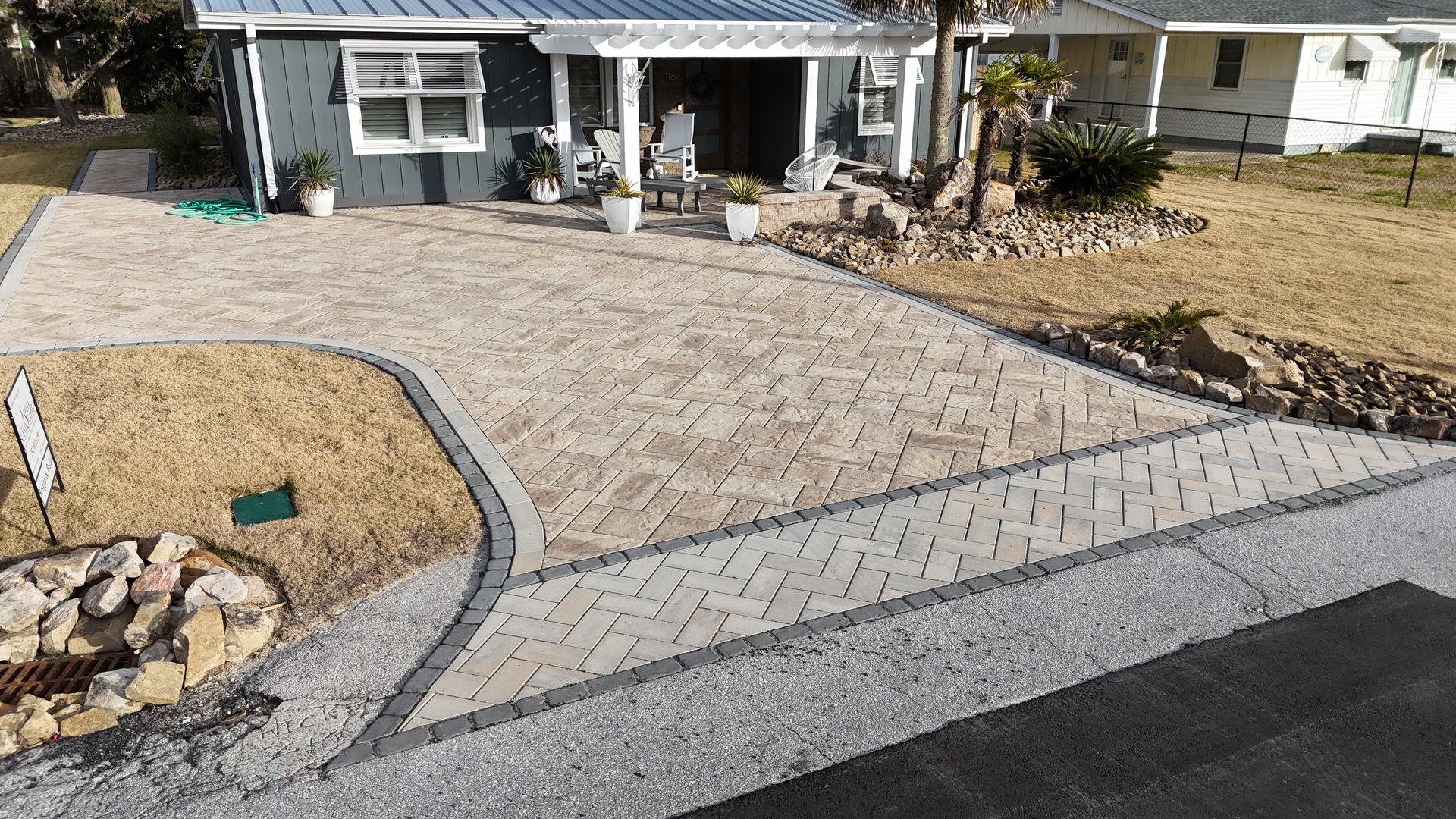 Brick paver driveway leading to a blue house with white trim; landscaping includes a small rock garden.