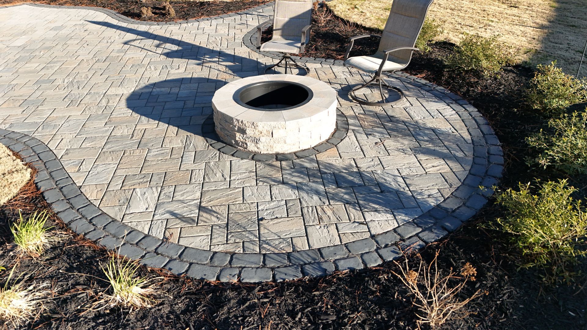 Stone patio with fire pit, bordered by dark pavers and landscaping.