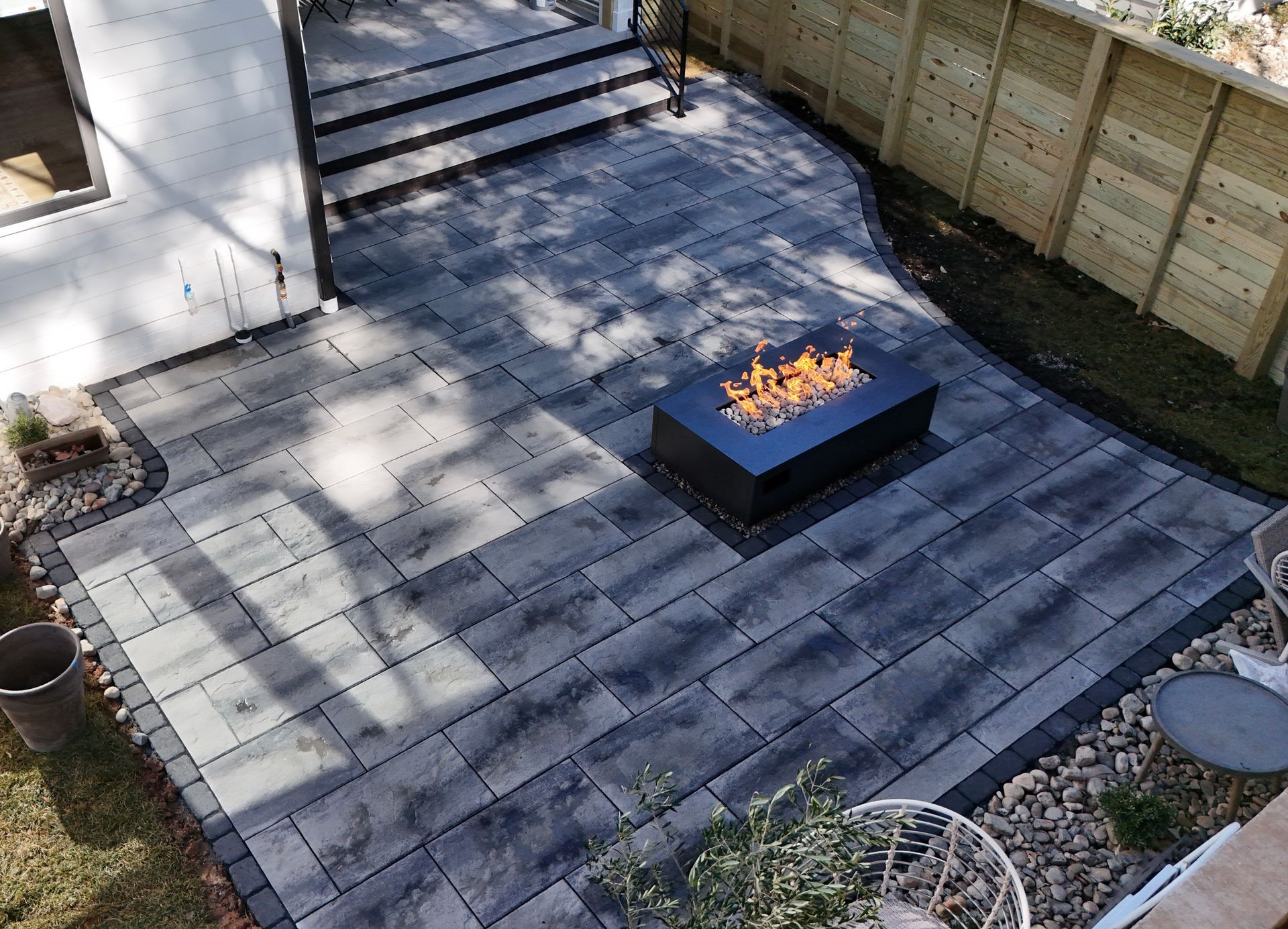 Patio with gray pavers, black fire pit, steps leading up, and a wooden fence.