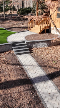 Brick pathway leading to wooden stairs and a house in a wooded area. Brown leaves and green lawn.