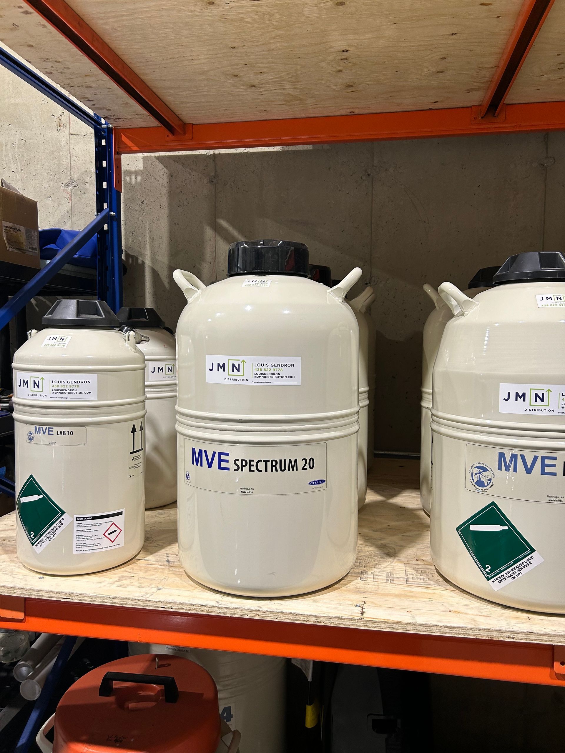 Several large, white cryogenic storage tanks with black lids and labels, on a shelf.