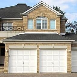 A two-story tan stone house with two white garage doors, a dark shingled roof, and a second-floor arched window.