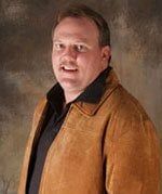 A person with a slight smile wears a brown suede jacket over a black collared shirt against a mottled brown background.