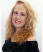 Portrait of a smiling person with shoulder-length, wavy blonde hair wearing a black off-the-shoulder top.