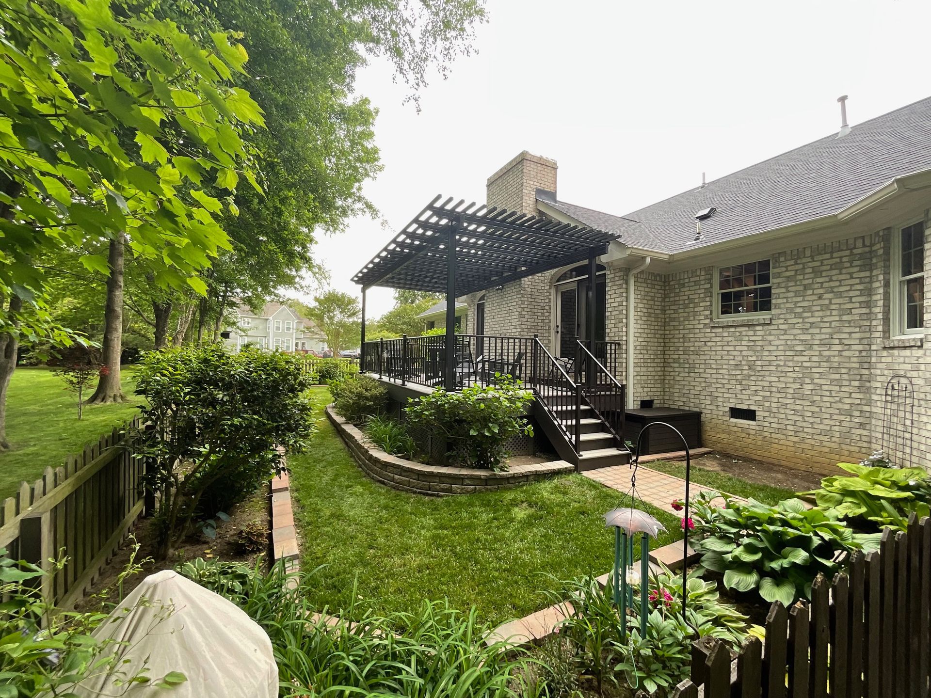 Backyard view showing a white brick house with a black wooden pergola over a deck, landscaped lawn, and wooden fencing.