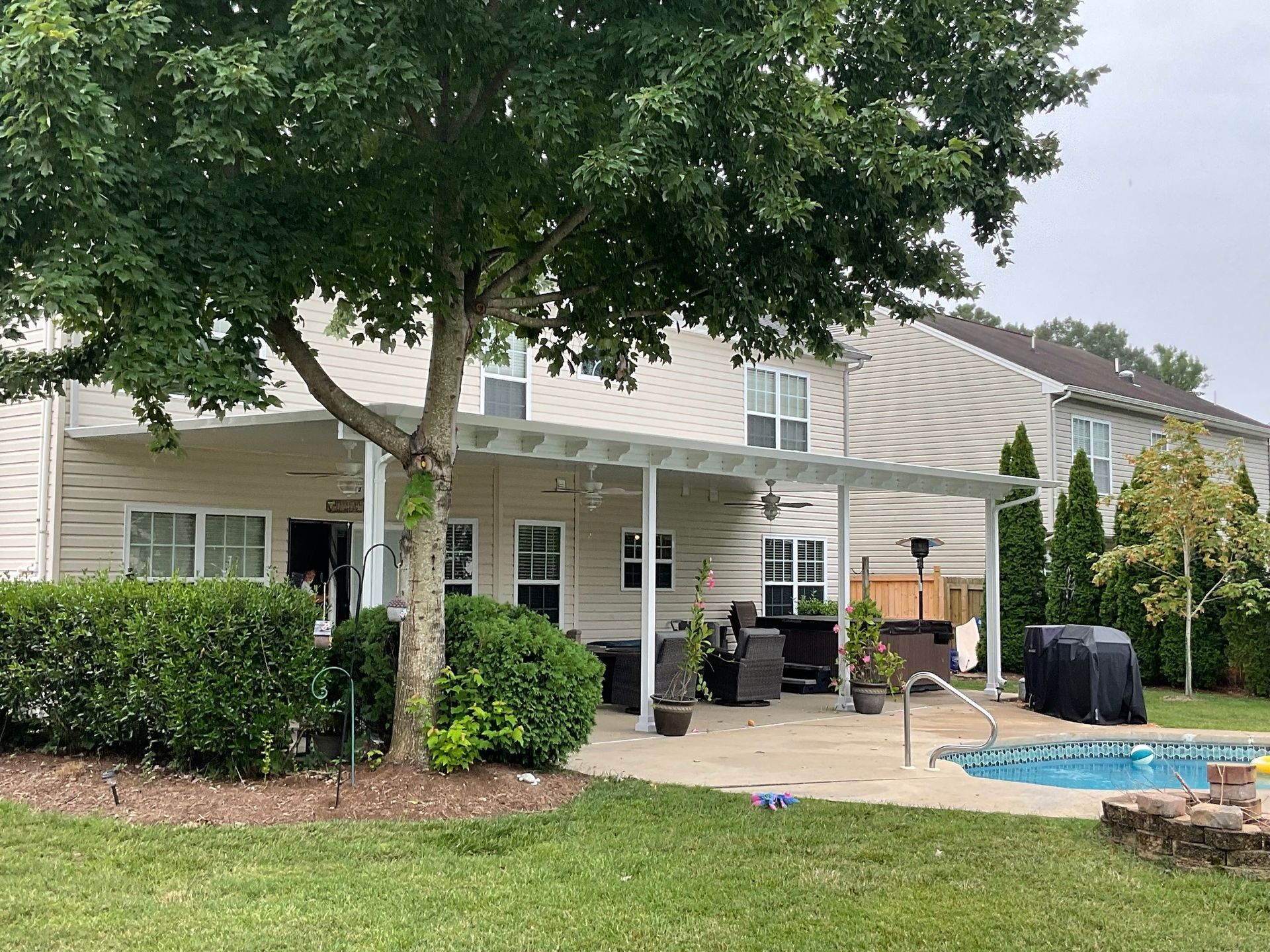 A backyard patio area featuring a white pergola, patio furniture, a swimming pool, and a large tree, attached to a house.