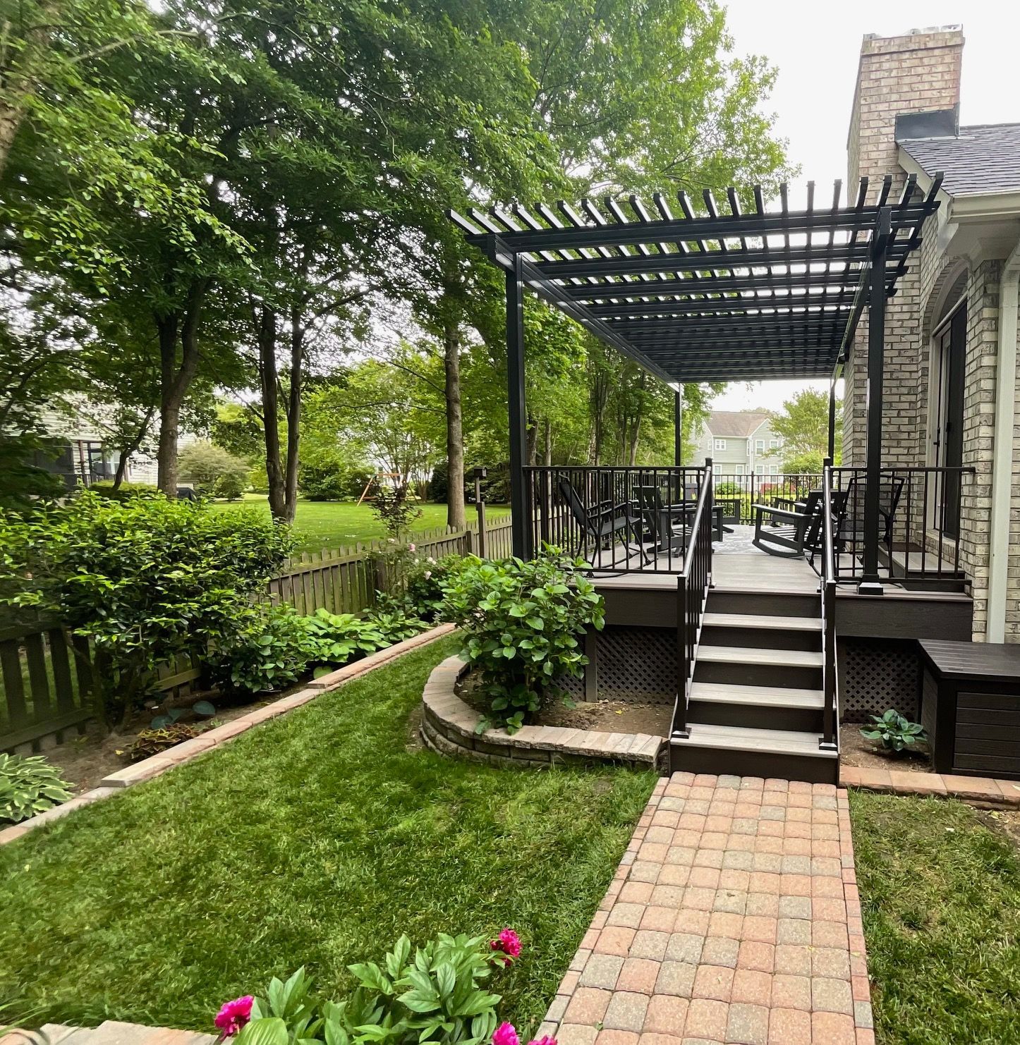 A brick walkway leads to a wooden deck with a black pergola and seating next to a stone house and a lush green backyard.