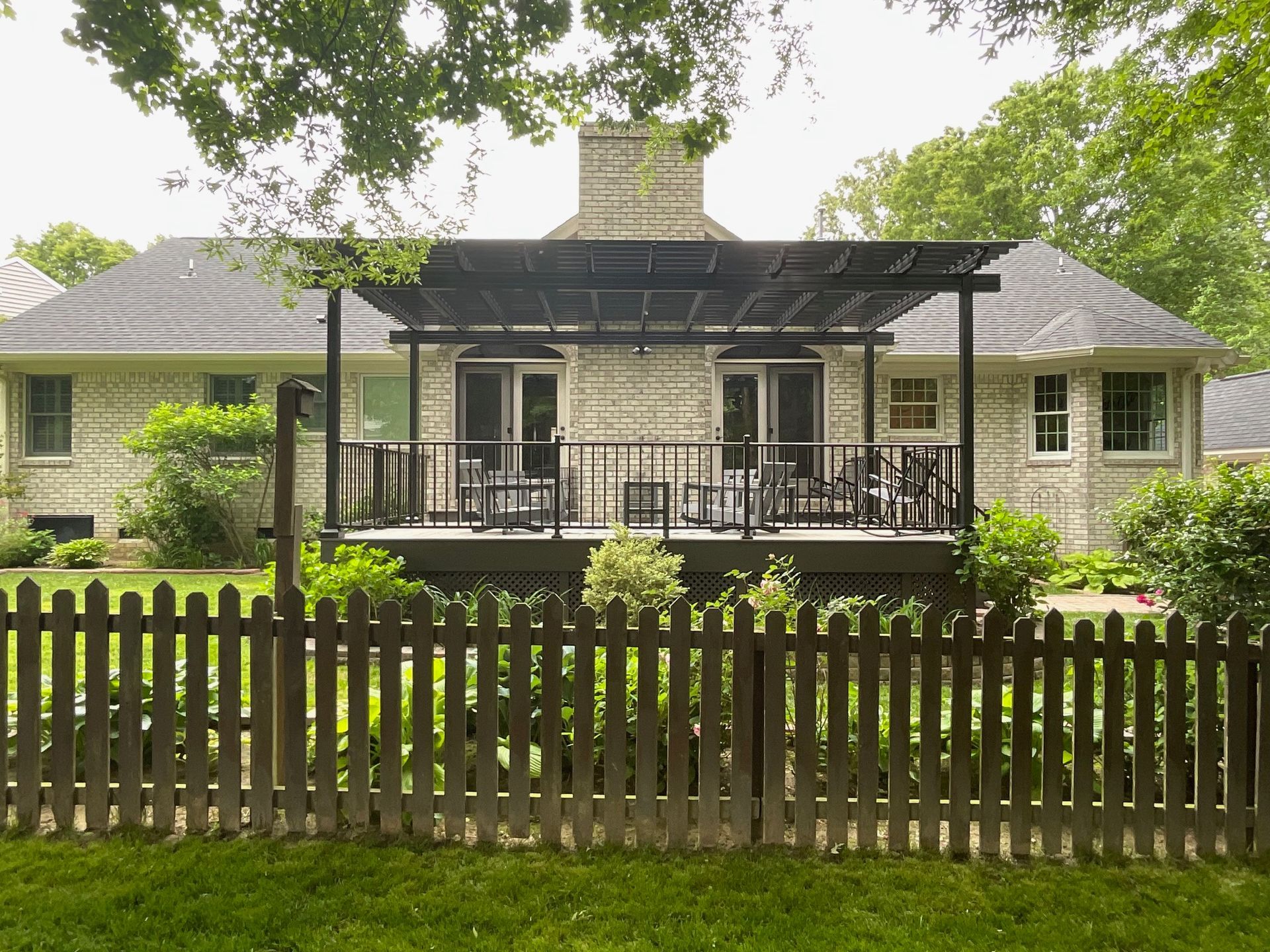 A light brick house with a dark pergola over a wooden deck, viewed from behind a wooden picket fence in a grassy yard.