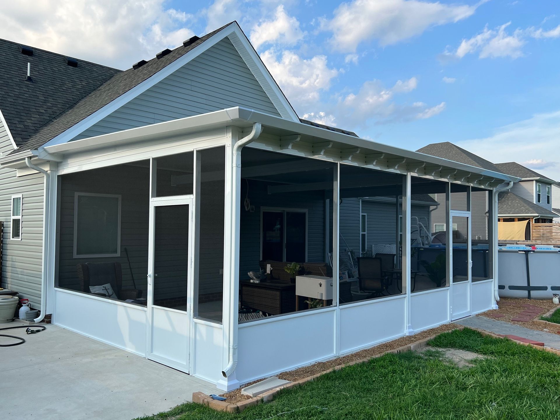 A white, screened-in porch addition attached to the side of a gray-sided suburban home with a concrete patio.