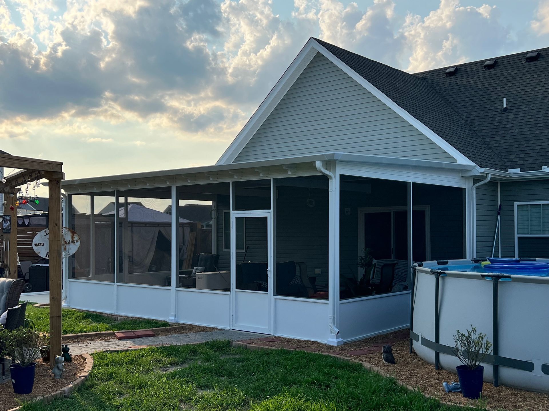 A white screened-in patio attached to the back of a house with a grey shingled roof, next to a circular above-ground pool.