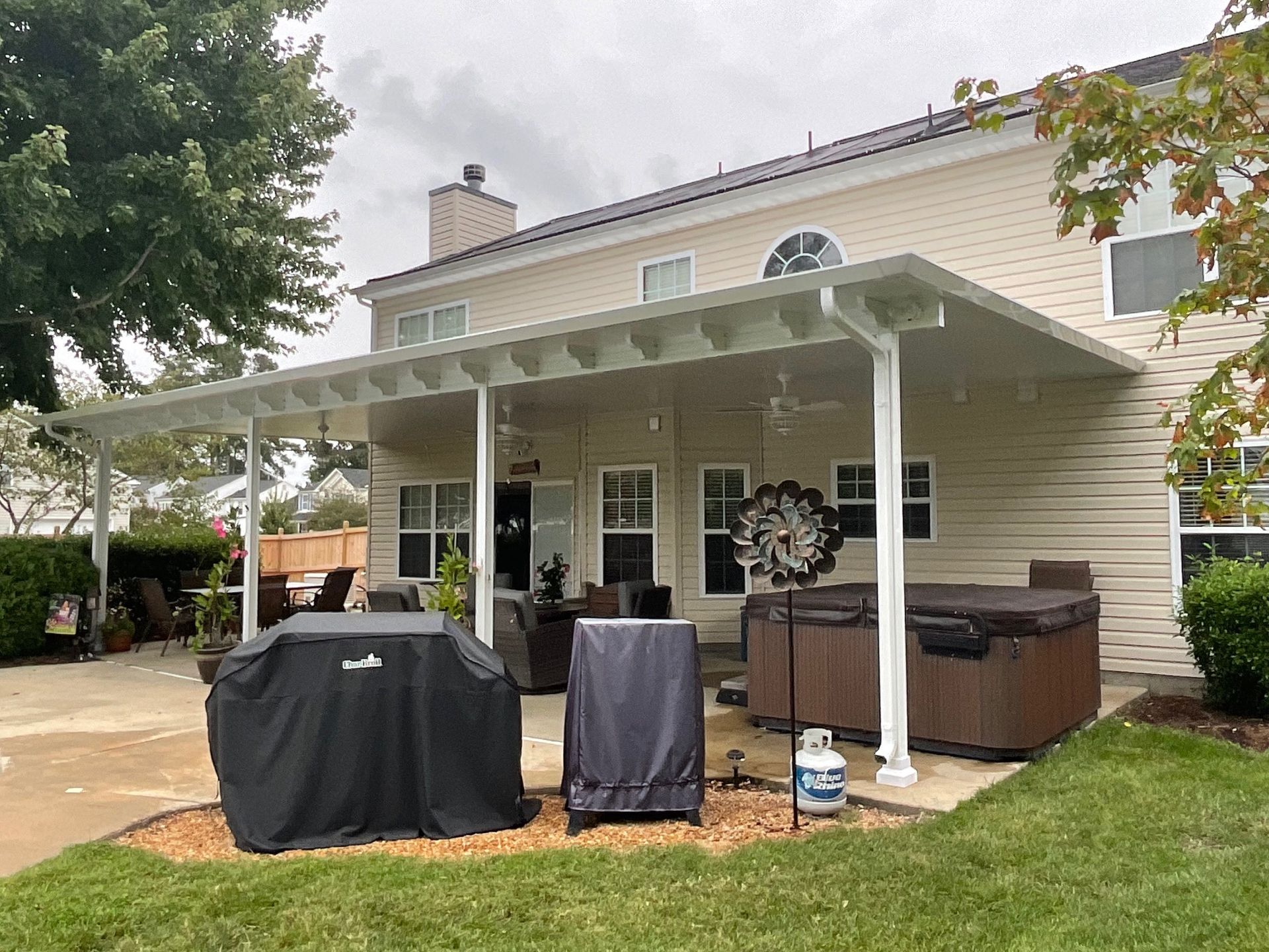 A tan house with a large covered patio featuring a grill, a smoker, and a hot tub on a concrete slab.