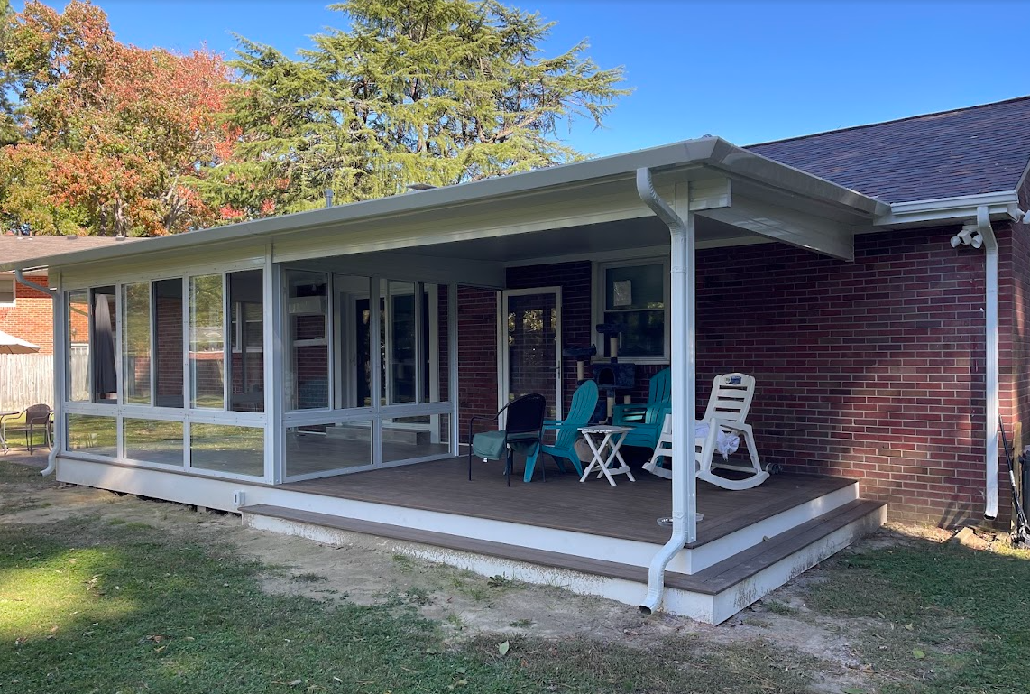 A brick house with a white-framed screened-in porch and an adjoining deck with blue and white chairs on a sunny day.