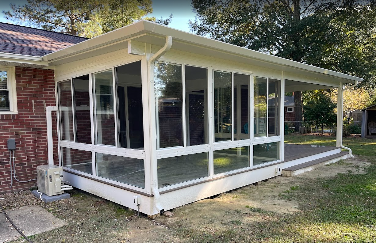 A white, window-enclosed sunroom attached to a brick house with a white roof and a side porch extension in a grassy yard.