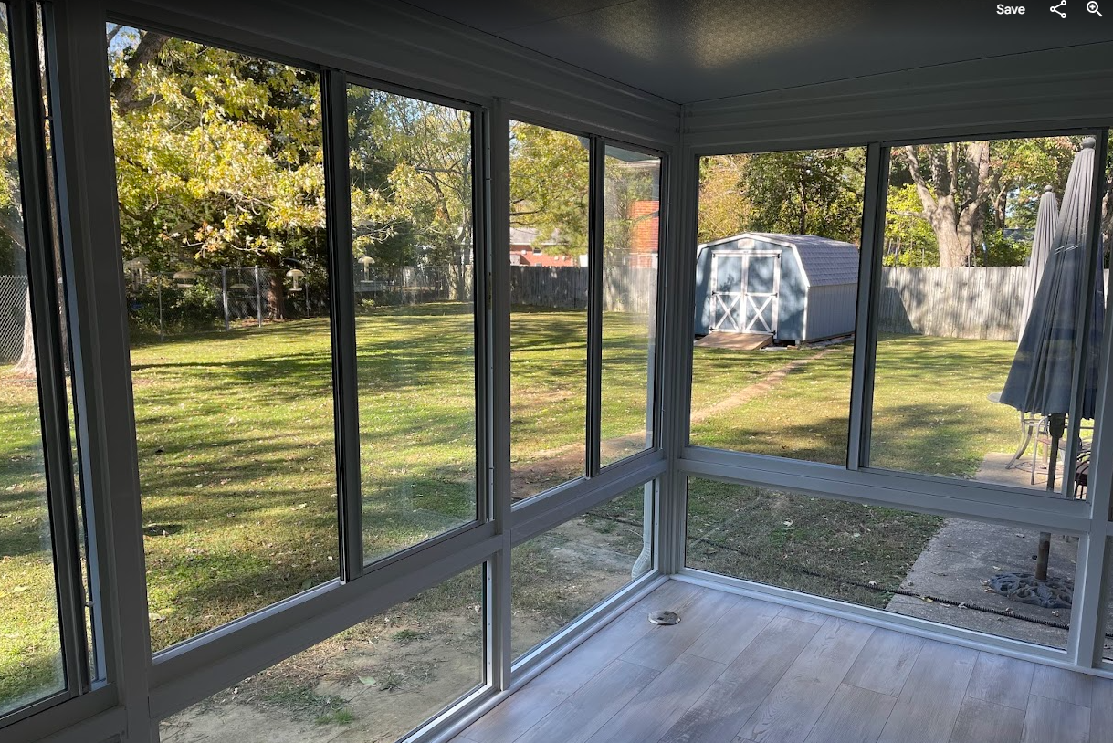 View from an enclosed sunroom looking out over a grassy backyard featuring a small storage shed and trees.