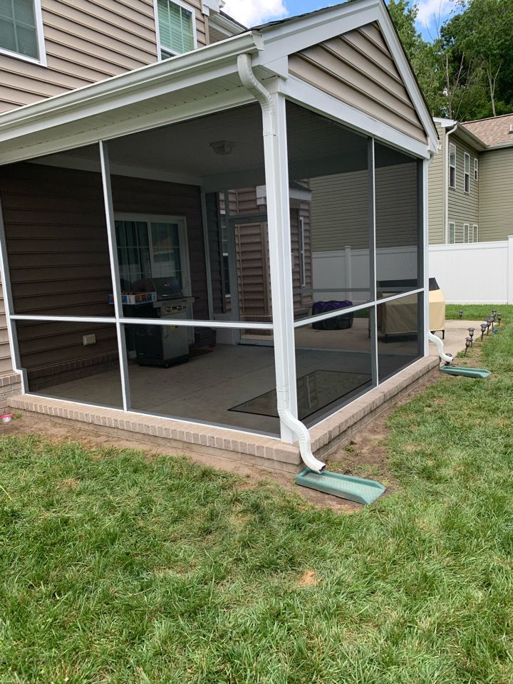 A screened-in porch with white framing, brick base, and attached downspouts, situated on a backyard lawn next to a house.