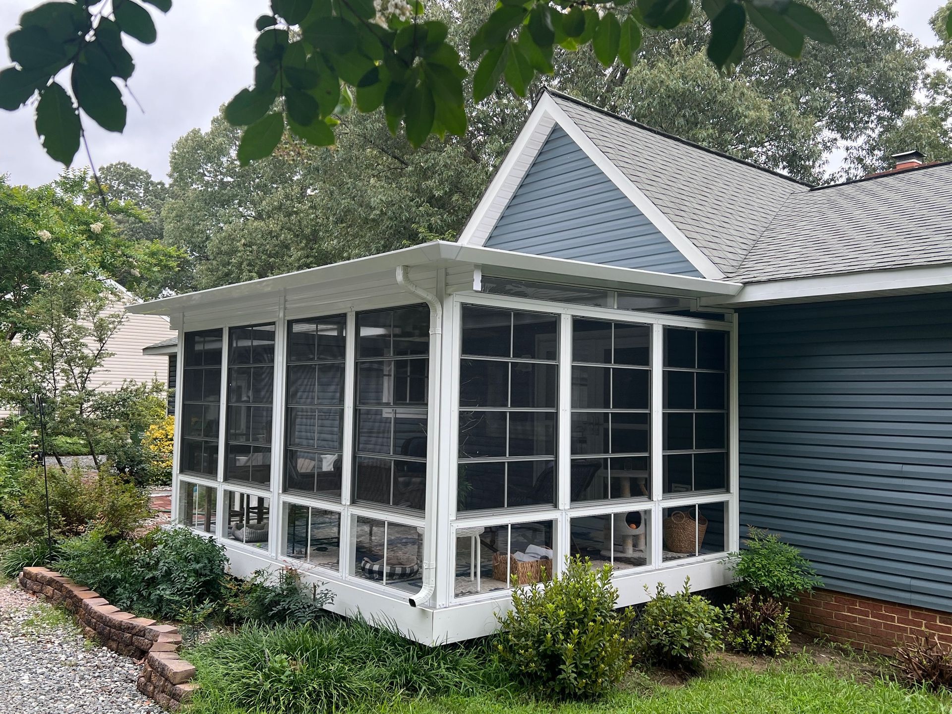 A screened-in porch with white frames attached to a blue-sided house, surrounded by trees and a brick-bordered garden.