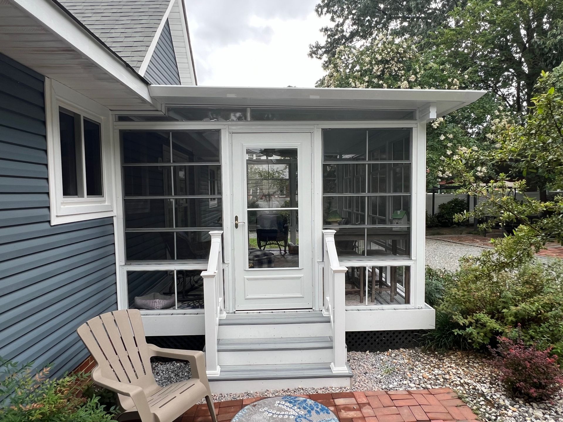 A gray-sided house exterior features a white-framed screened-in porch with a central door and steps leading to a patio.