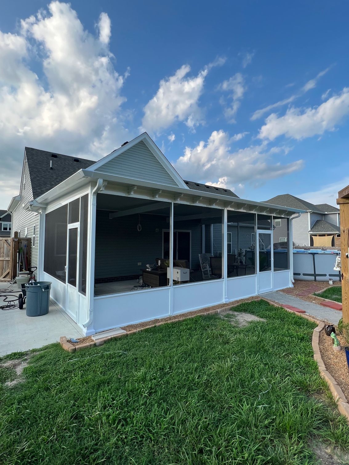 A white screened-in porch attached to the back of a house under a partly cloudy blue sky, next to a grassy lawn.