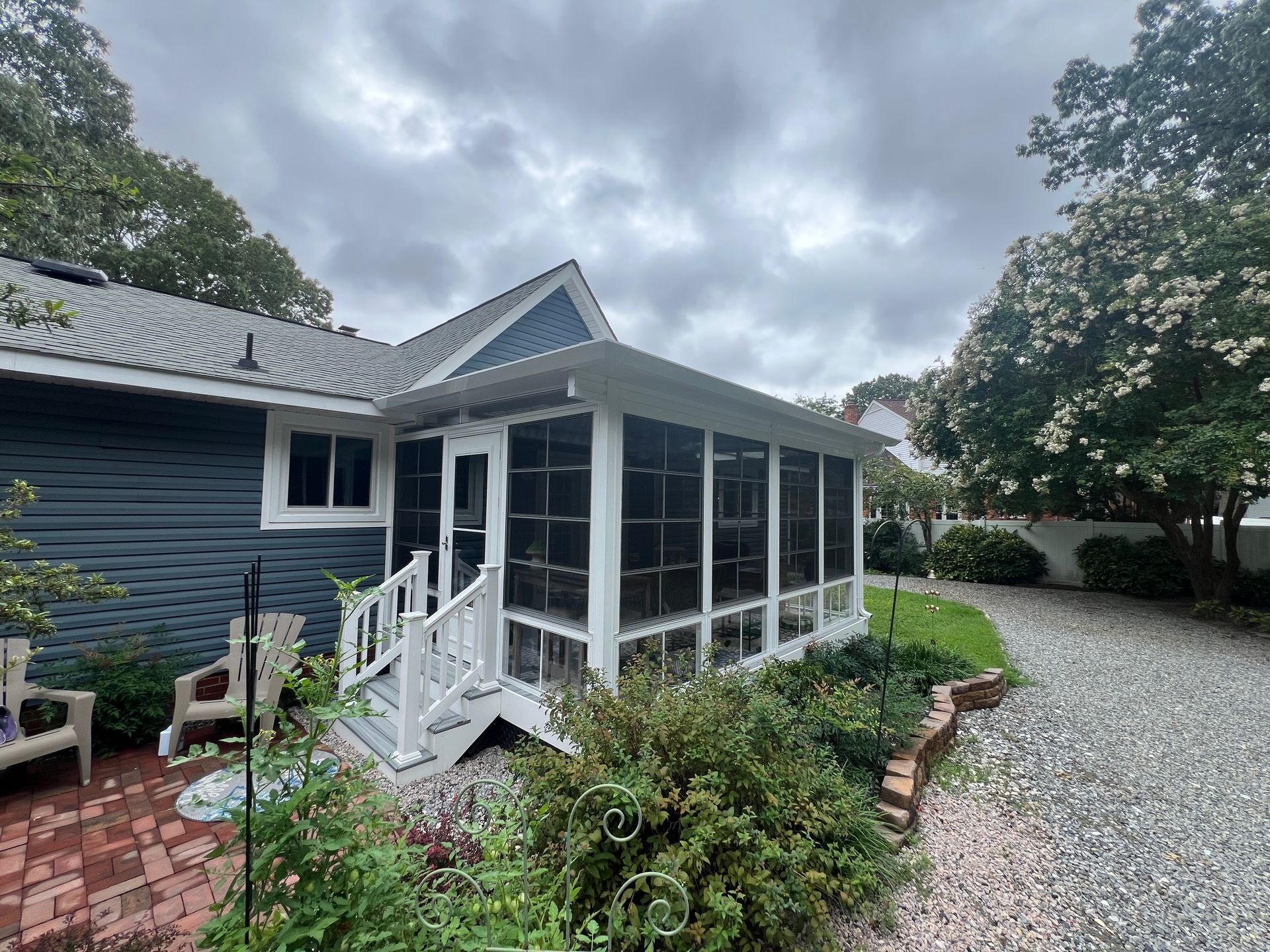 A blue house with a white screened-in porch, brick patio, and gravel driveway under a cloudy sky.