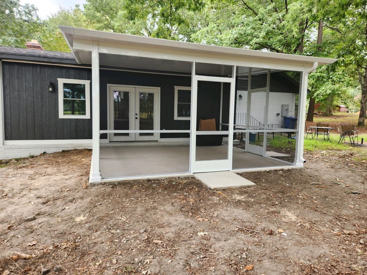 A modern white screened-in patio with a shed-style roof attached to the back of a dark-painted house with a dirt yard.