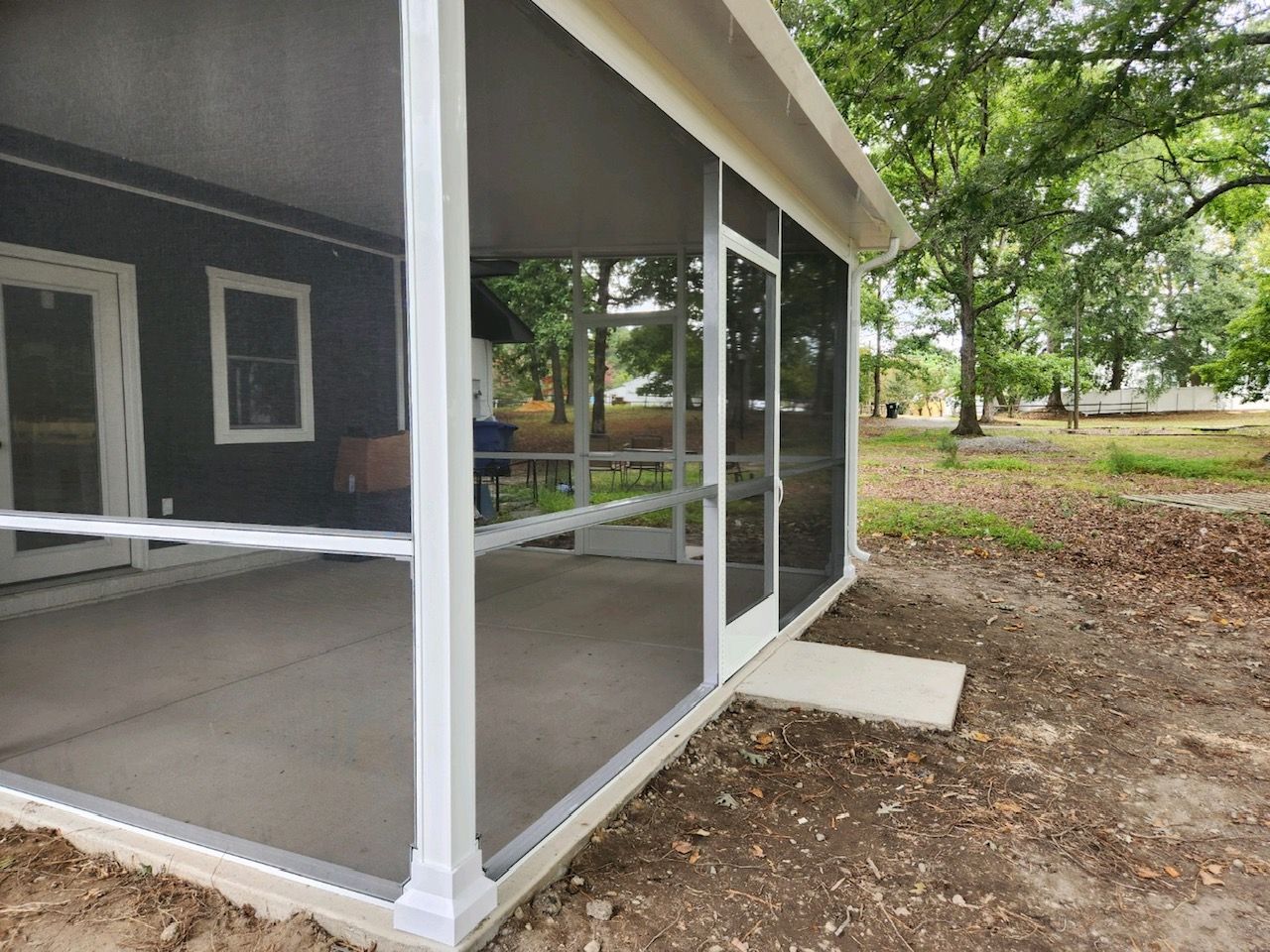 A screened-in porch with white framing and concrete flooring, positioned next to a dark gray house in a wooded area.