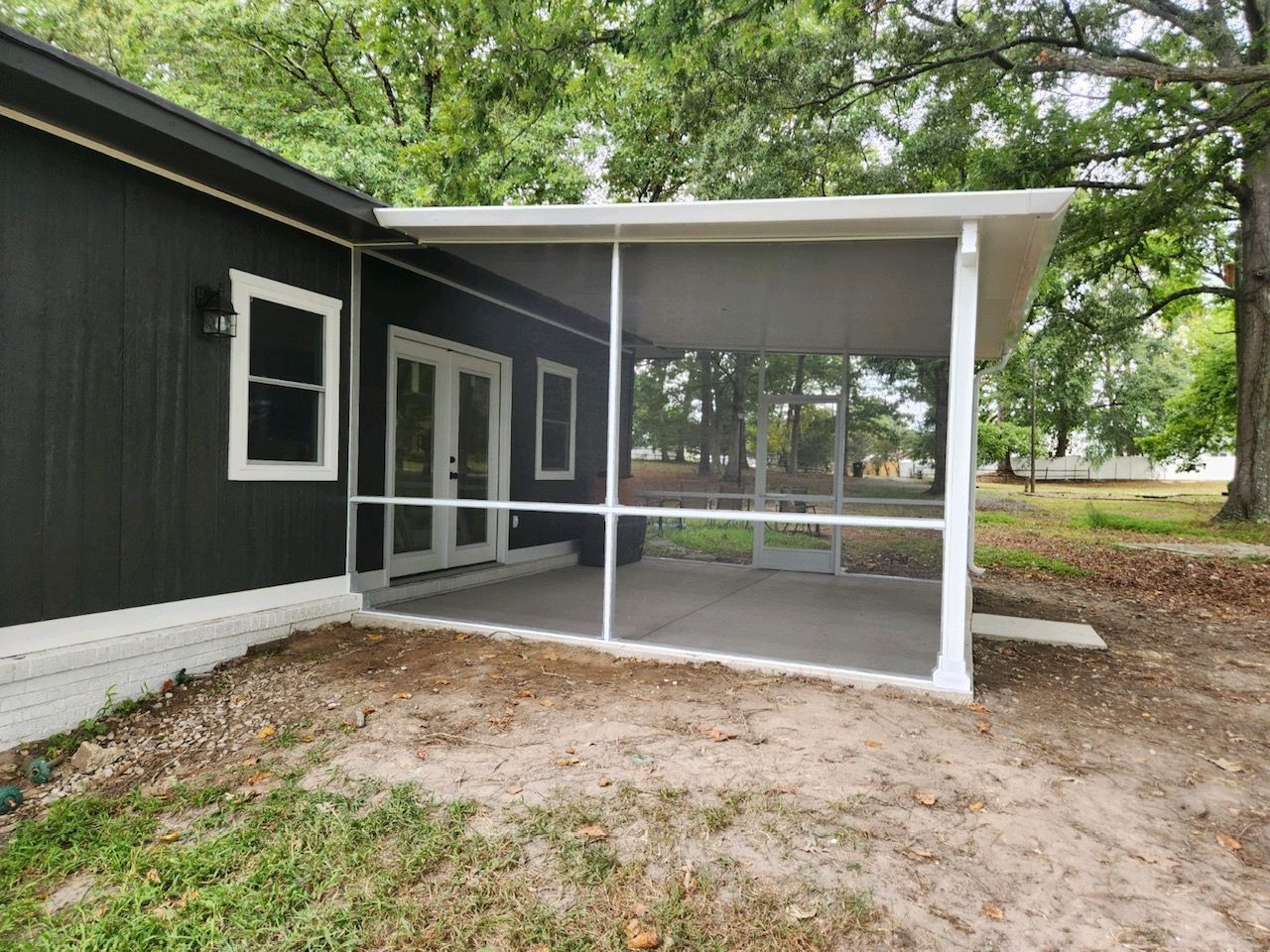 A dark-sided house with a white-framed screened-in porch, glass double doors, and a concrete floor in a wooded yard.