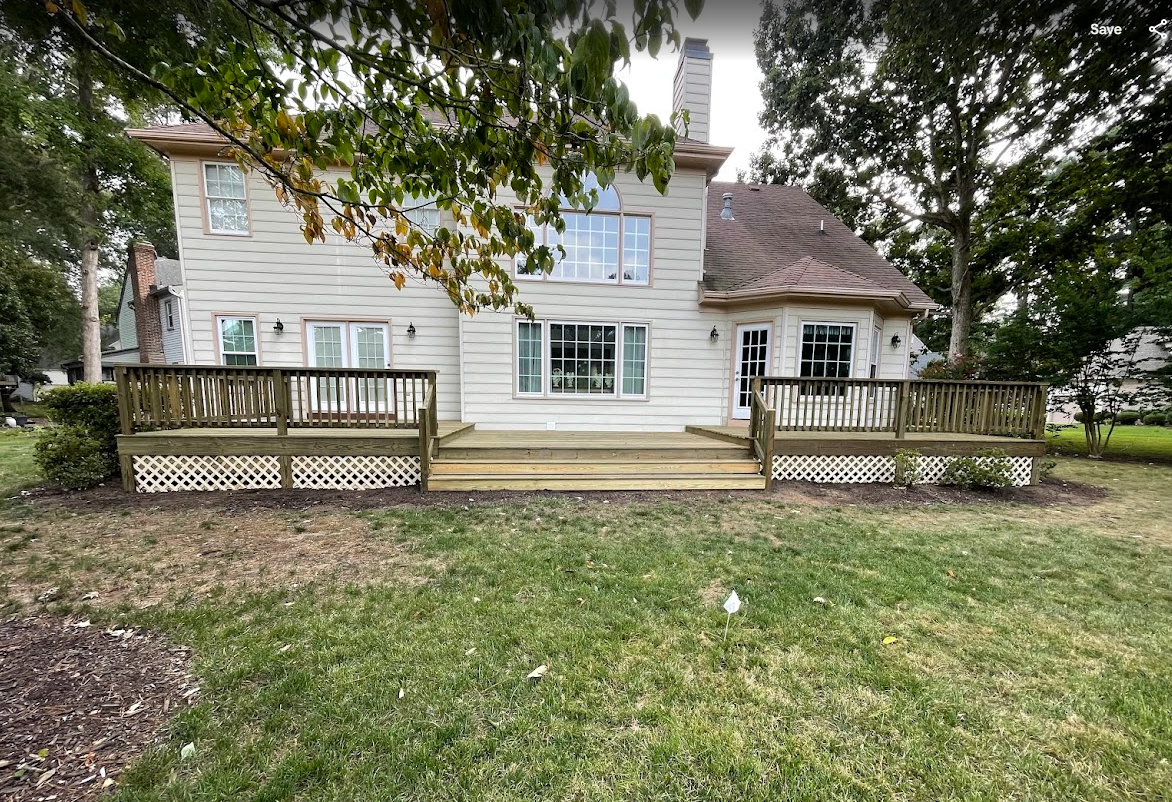 A two-story house with a large wooden deck, white siding, and a green lawn under a leafy tree.