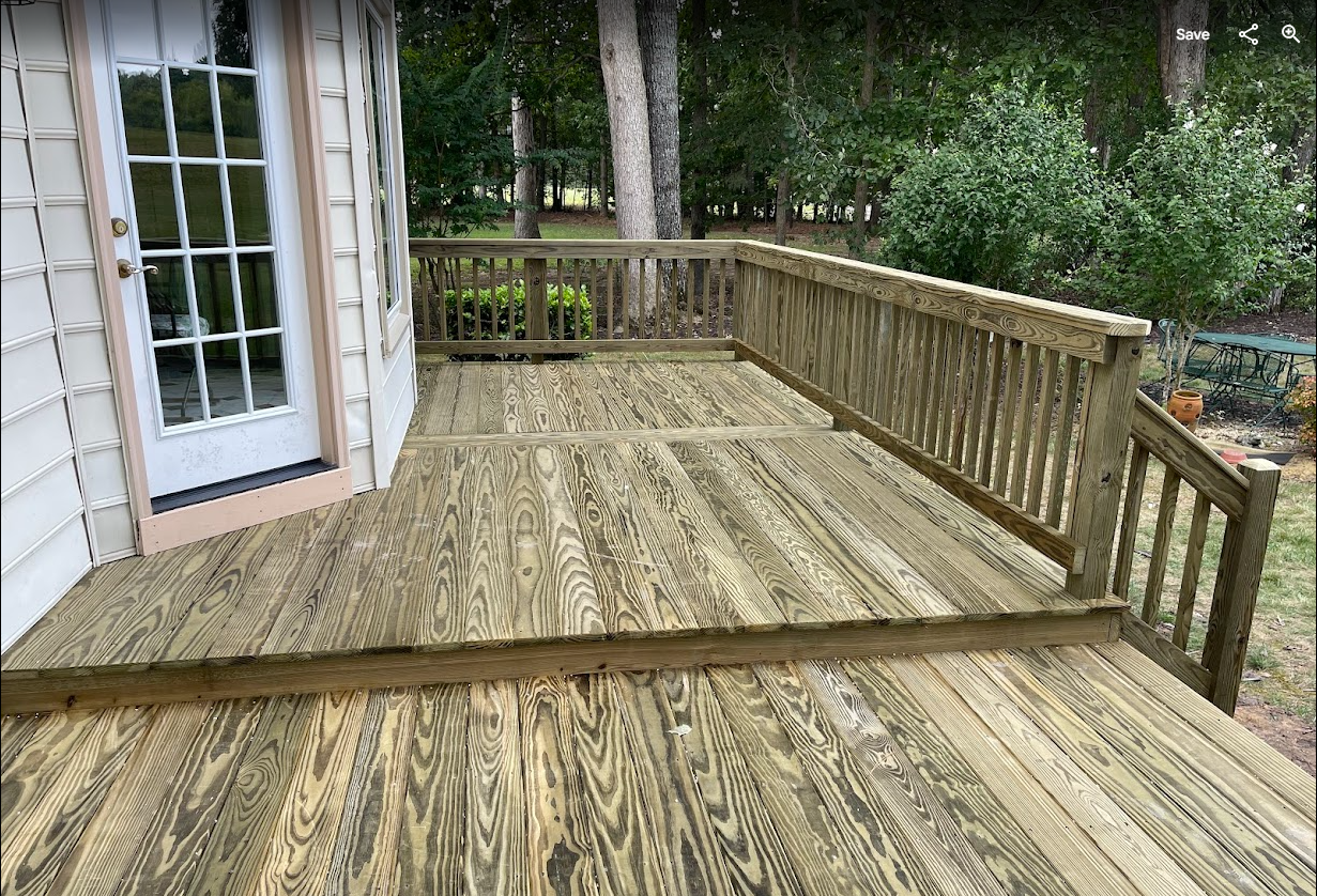A two-level wooden deck attached to a house with a white door and wood railing surrounded by trees.