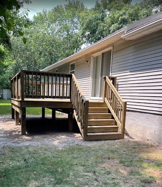 A wooden deck with stairs attached to the back of a house with light gray siding and a glass sliding door.