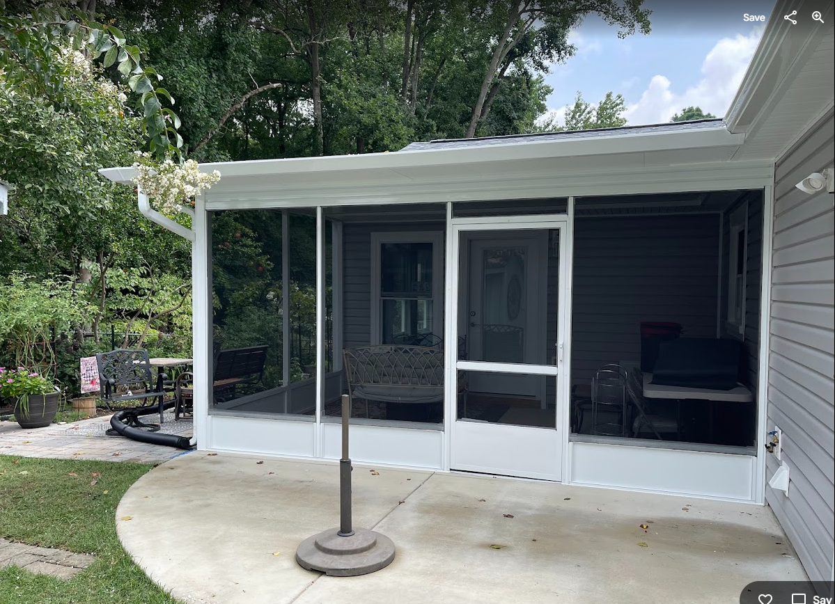 A screened-in porch with a white frame attached to a gray house, featuring a concrete patio and a patio umbrella stand.