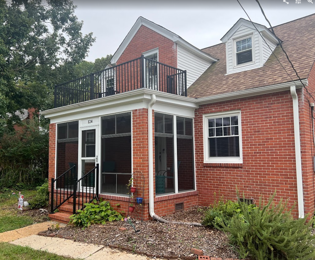 A two-story brick house with a screened-in porch, an upper deck with a black railing, and a dormer window on the roof.