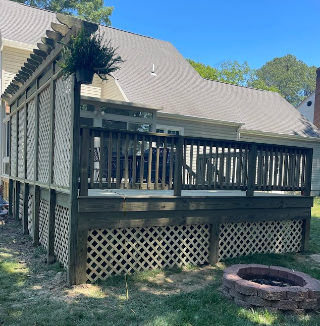 A dark-stained wooden deck with a trellis and lattice skirting sits in a backyard next to a house with a stone fire pit.