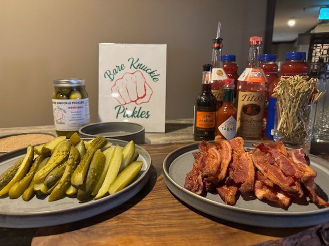 Platters of pickles and bacon, Bloody Mary fixings, and Bare Knuckle Pickles sign.