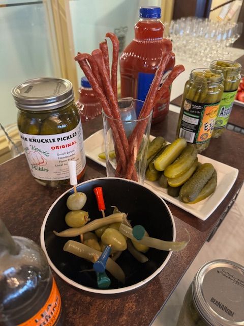 Pickles, olives, meat sticks, and Bloody Mary mix arranged on a table for appetizers.