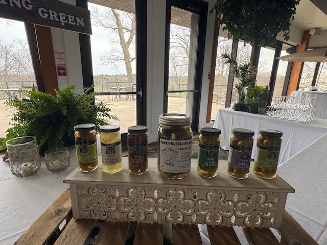 Jars of pickles and relishes on a decorative white wooden tray displayed indoors with a window view of trees.