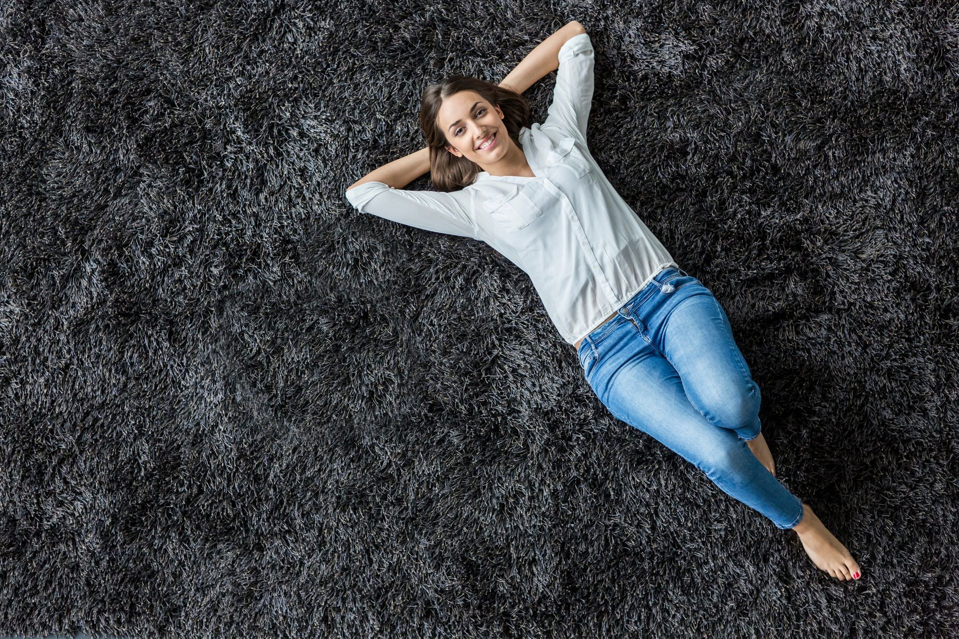 Woman with hands behind her head lying on a soft, dark gray carpet and smiling.