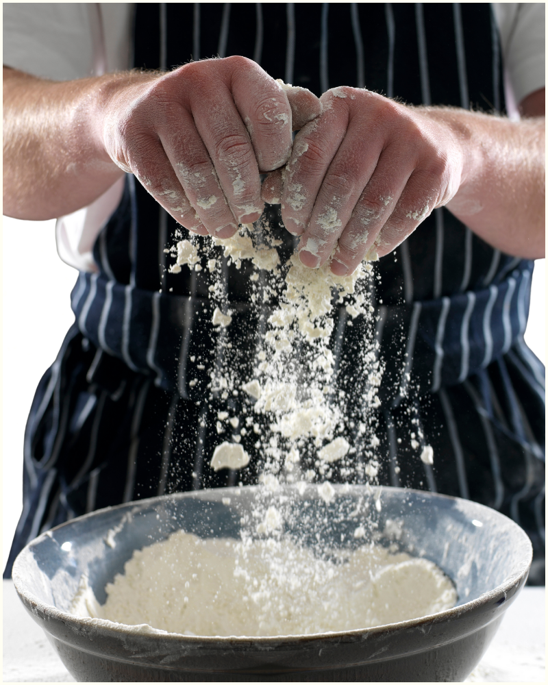 A person in a striped apron dusting flour through their fingers into a large, rustic bowl.