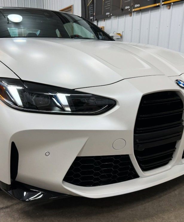 A close-up of a matte white BMW M4 front end, highlighting its signature large grille and angular LED headlights.