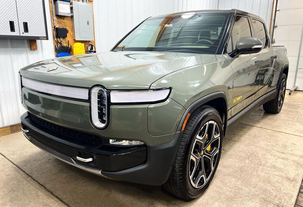 Olive green Rivian electric truck parked indoors with shiny surface, black trim, and yellow accents.